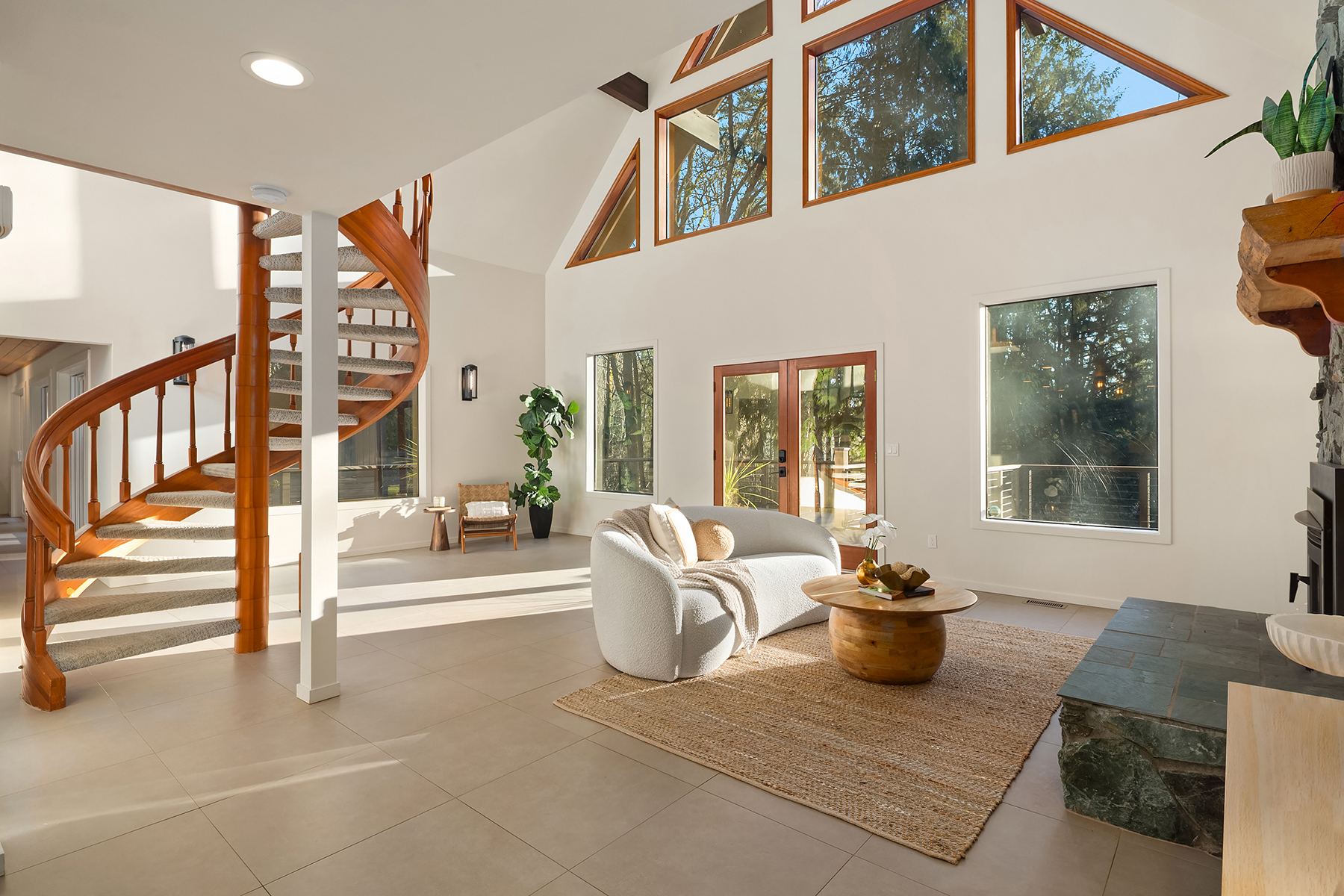 The vaulted great room with peaked window wall, exposed beams, spiral staircase, and stone hearth